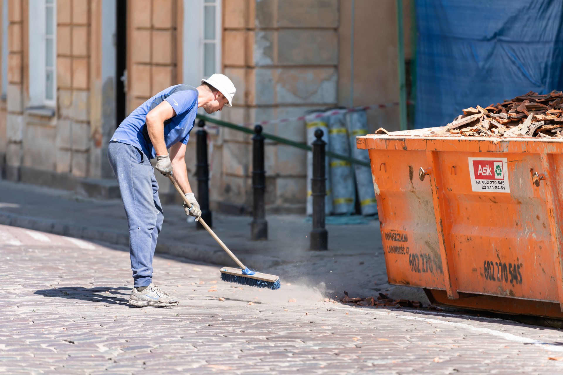 Old or new town street with man worker cleaner in blue uniform cleaning trash dust from cobblestone cobbled road by dumpster and sweeper tool during day